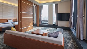 Habitación de hotel moderna con cama, sofá, televisor y grandes ventanales que ofrecen vistas a la ciudad.