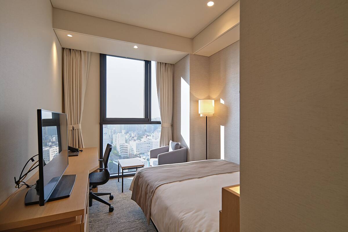 Une chambre d’hôtel équipée d’un lit, d’un bureau, d’une chaise et d’une grande fenêtre donnant sur la ville.