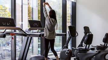 Un gimnasio con cinta de correr, bicicletas estáticas y un ventanal con vistas a árboles y edificios.