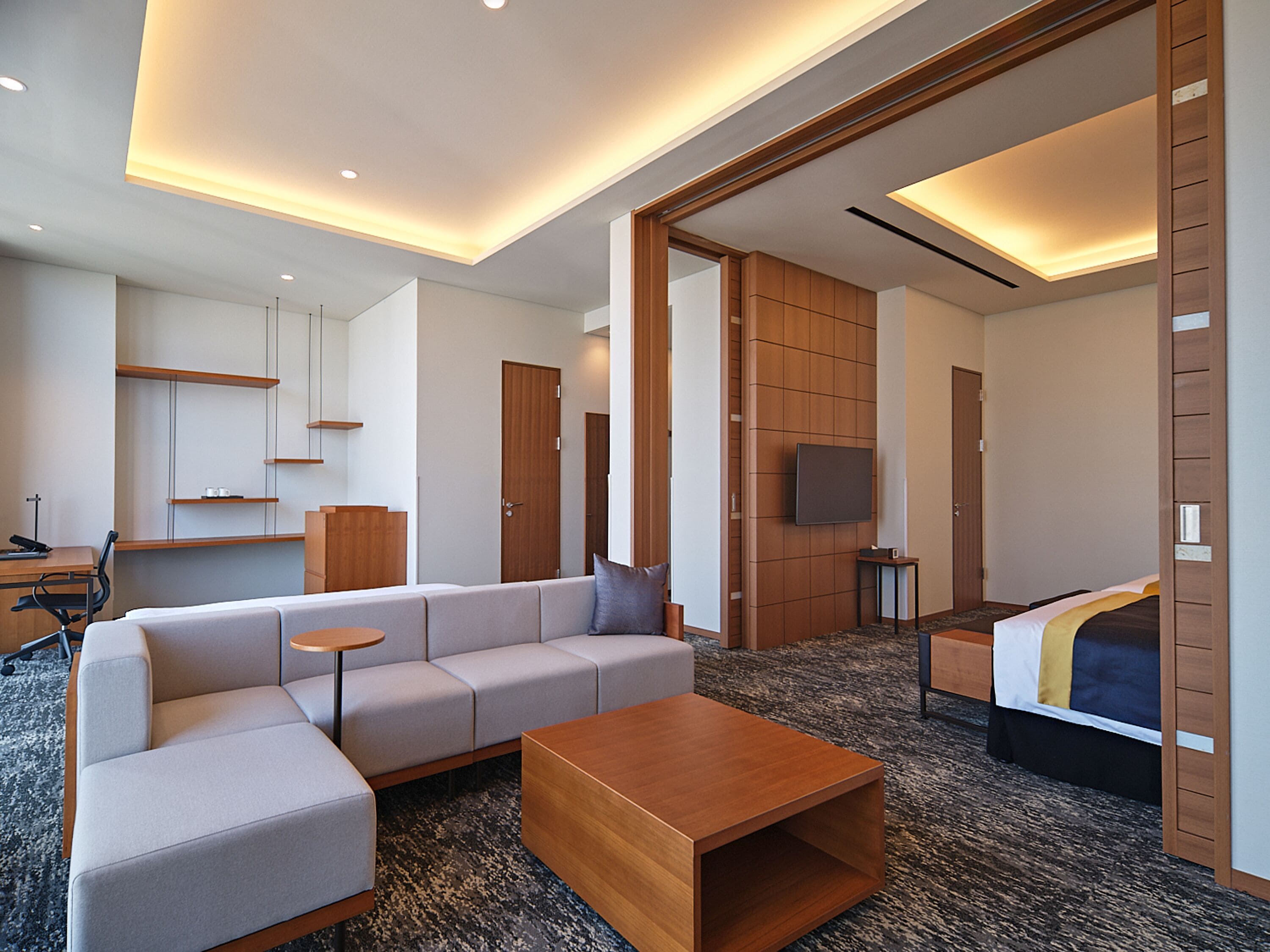 A modern hotel room with a sofa, a wooden coffee table, a desk with a chair, a TV, and a bed with a blanket.