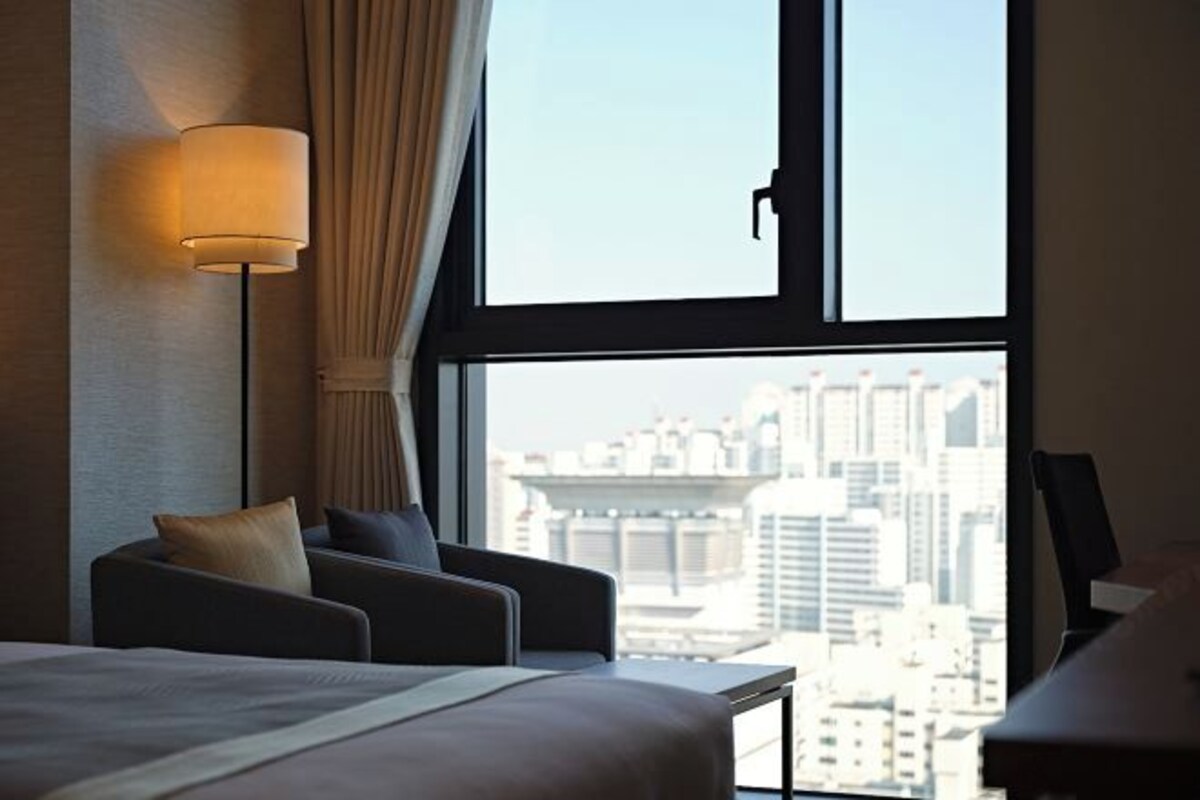 A hotel room with a bed, two armchairs, a desk, a lamp, and a large window with a city view.