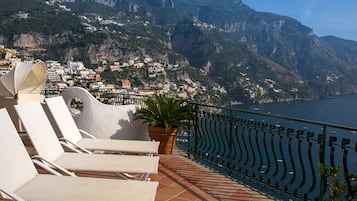 Une terrasse avec des fauteuils de jardin blancs et un pot de fleurs, d’où l’on peut admirer une ville côtière avec des bâtiments construits sur une colline et la mer en arrière-plan.