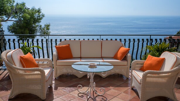 Ein Balkon mit Korbmöbeln, einem Glastisch und Blick aufs Meer.