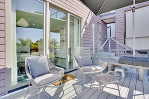 Eine überdachte Terrasse mit weißen Korbstühlen, einem Tisch, großen Schiebeglastüren und einer Treppe zu einer oberen Ebene.