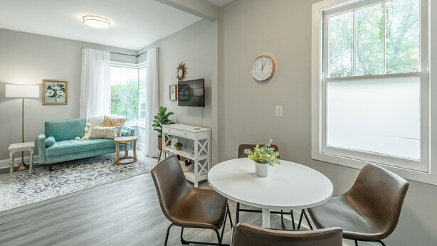 Una sala de estar con un sofá color turquesa, una mesa redonda blanca y sillas de cuero marrón. Hay un televisor sobre un mueble blanco, un reloj en la pared y una ventana con vistas a árboles.