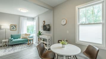 Un salon avec un canapé de couleur turquoise, une table ronde blanche et des fauteuils en cuir marron. On y trouve un téléviseur posé sur un meuble blanc, une horloge au mur et une fenêtre donnant sur des arbres.