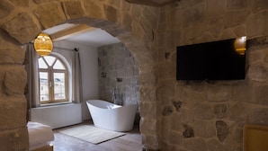 A stone-walled bathroom with a freestanding bathtub, a large window, and a flat-screen TV mounted on the wall.