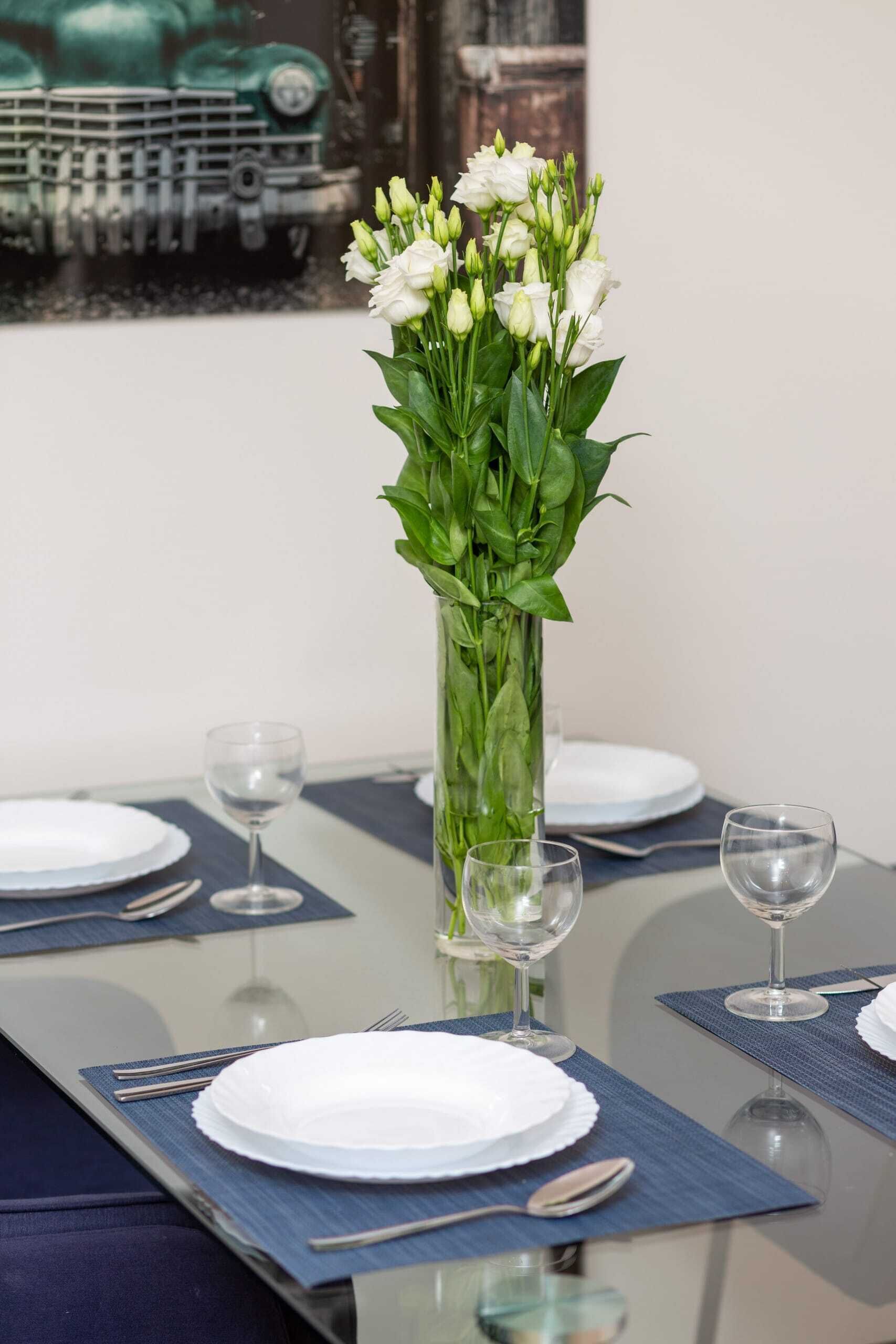 Uma mesa de jantar posta com pratos brancos, talheres e copos de vinho, com um vaso alto de flores brancas no centro.