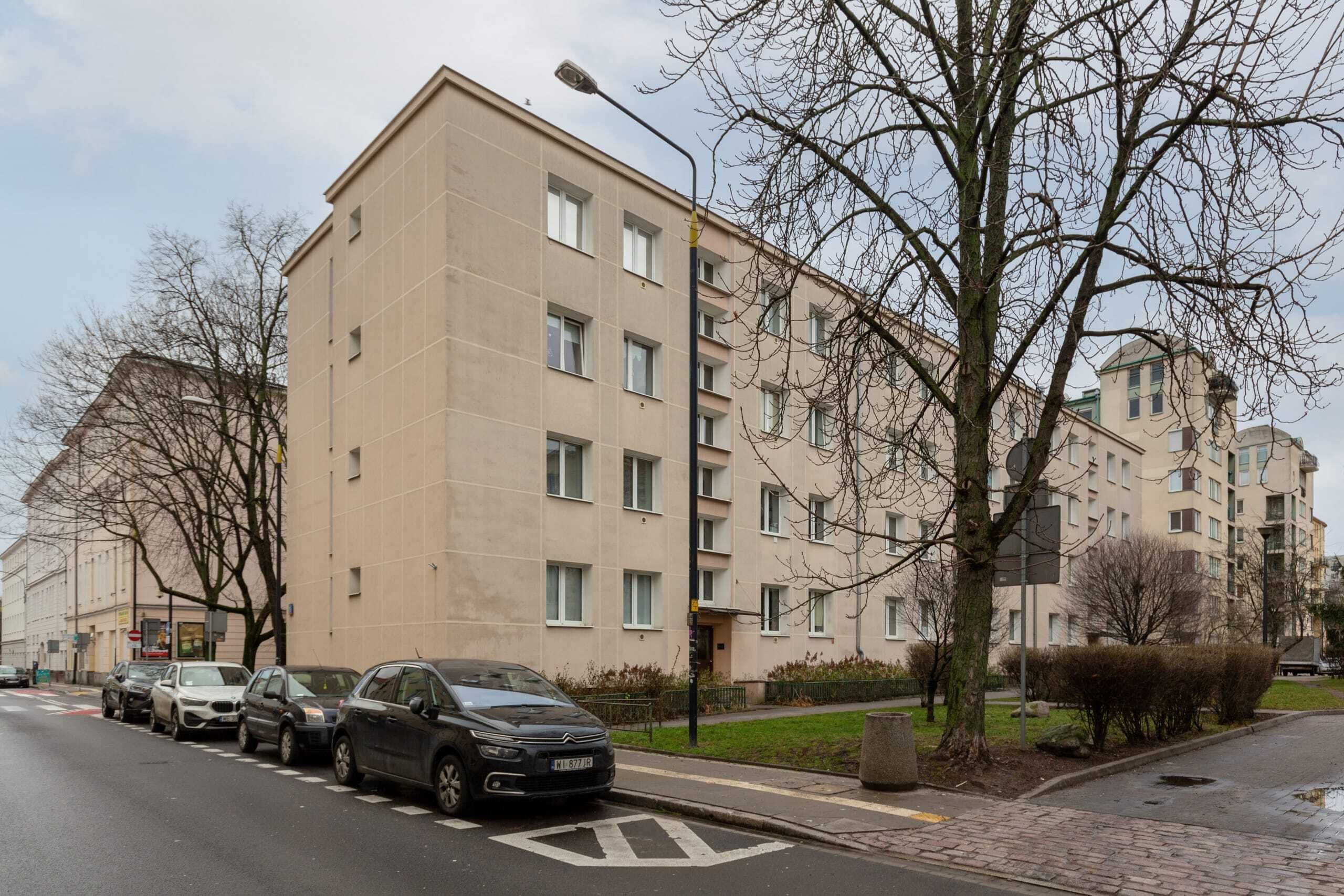 A multi-story residential building with a beige facade, parked cars along the street, and leafless trees.