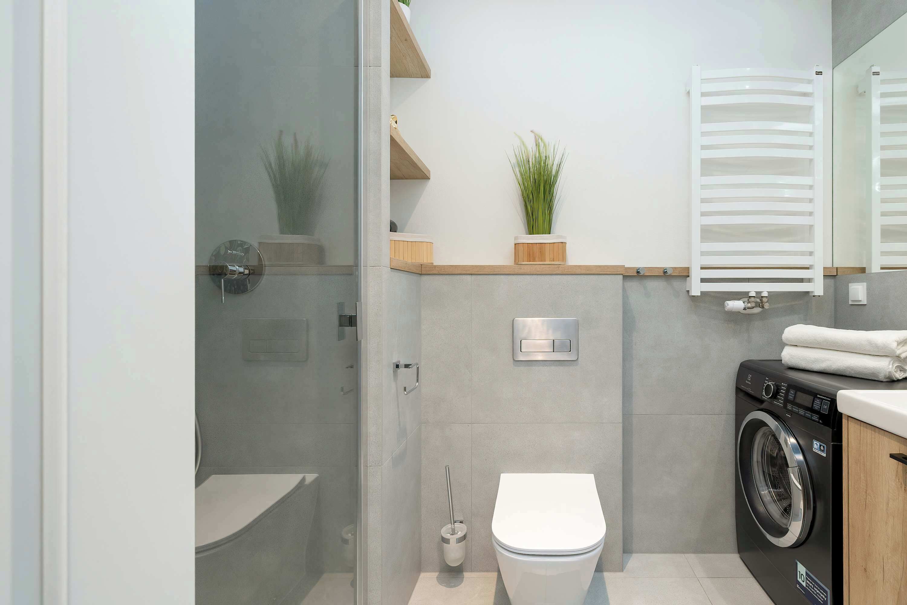 A modern bathroom with a glass shower enclosure, a toilet, a washing machine, and wooden shelves with plants.