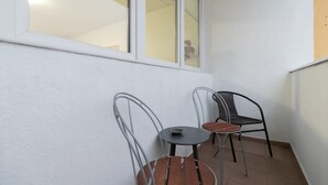 A small balcony with two metal chairs and a small table, white walls, and a window.
