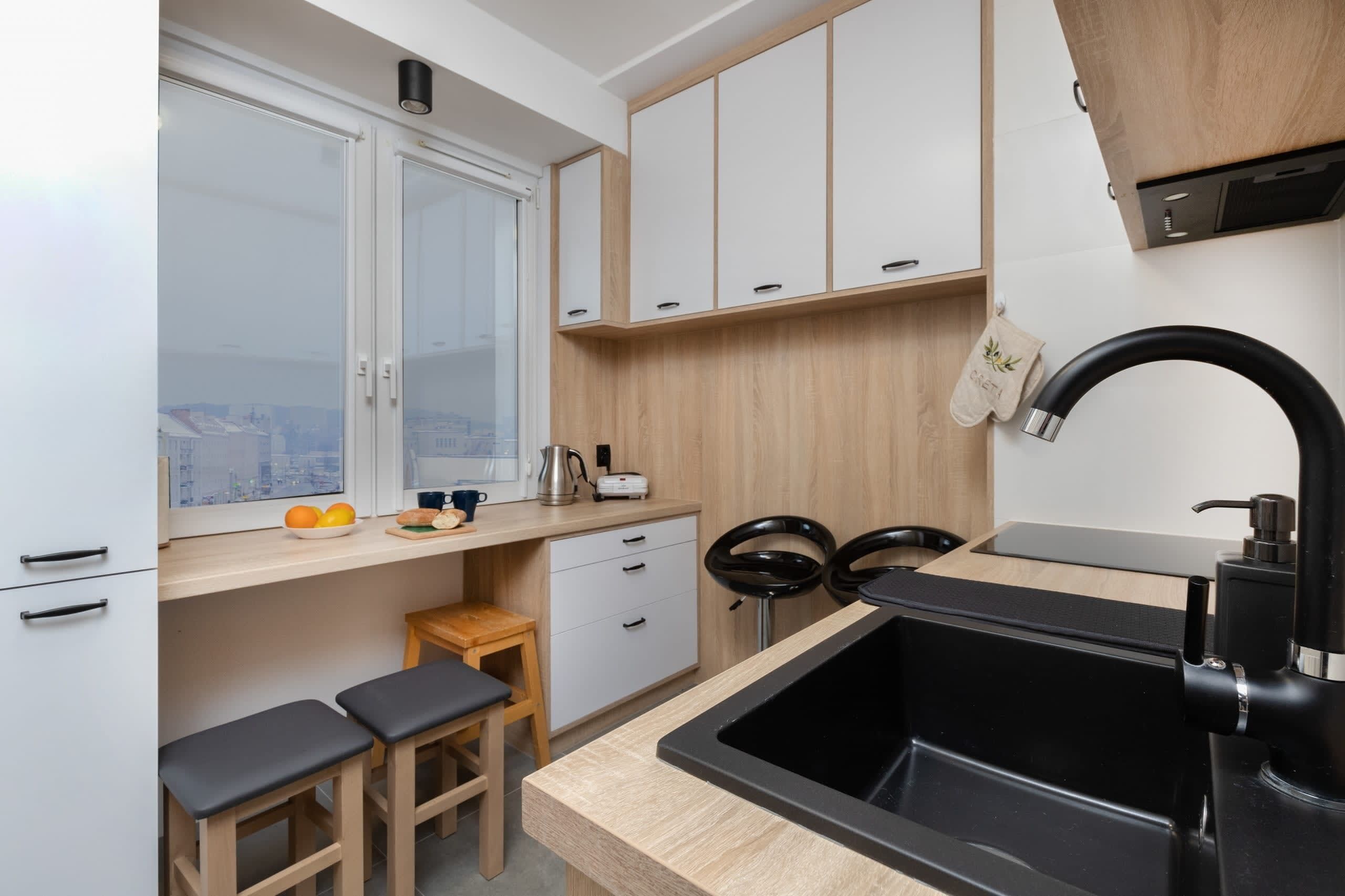 A modern kitchen with a black sink, wooden cabinets, a window with a city view, and a small stool.