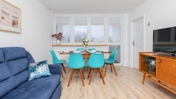 A modern living room with a blue sofa, teal chairs, a wooden dining table, and a TV on a wooden cabinet.