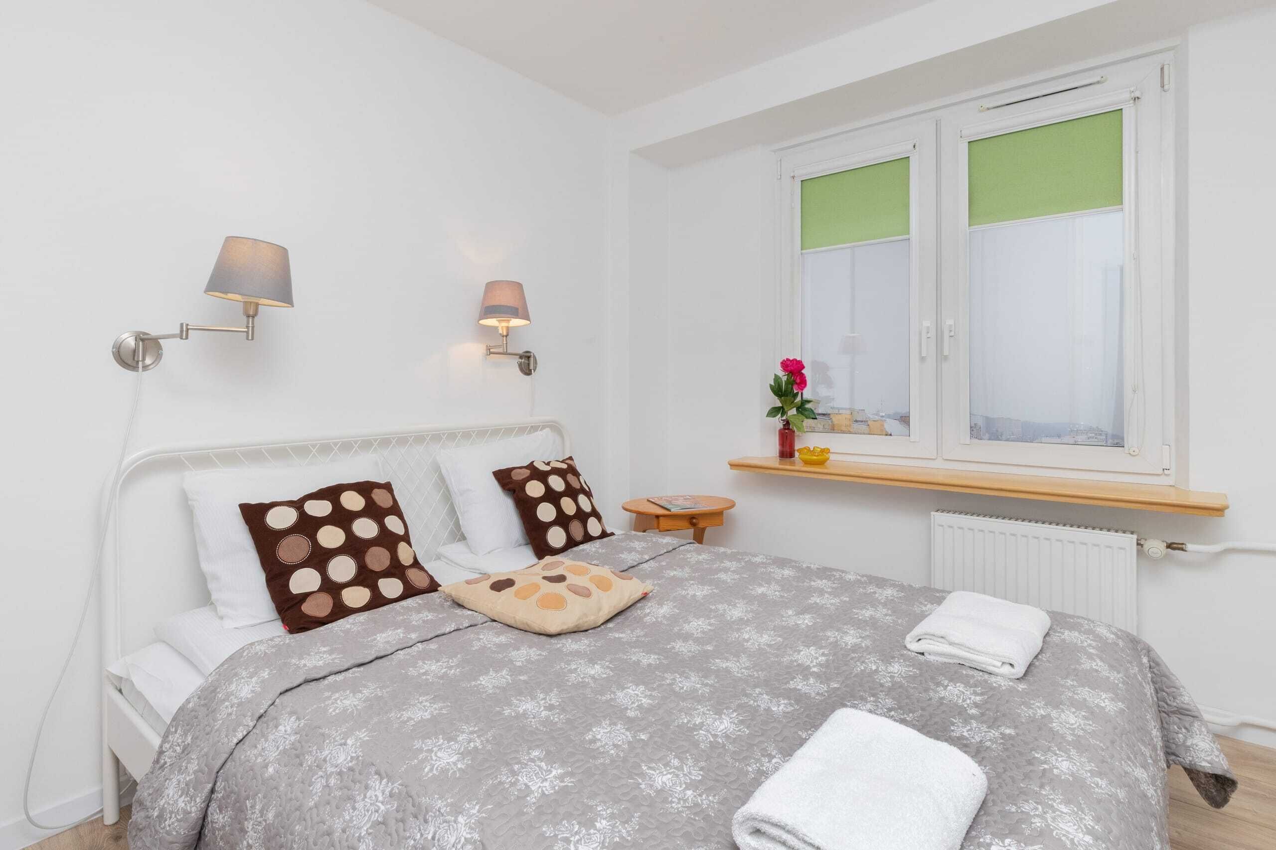 A neatly made bed with a grey floral bedspread, two wall-mounted lamps, a window with green blinds, and a small wooden nightstand with a vase of flowers.