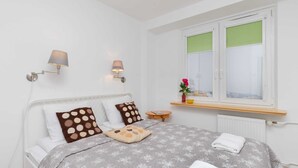 A neatly made bed with a grey floral bedspread, two wall-mounted lamps, a window with green blinds, and a small wooden nightstand with a vase of flowers.