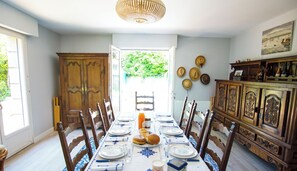 A dining room with a long table set for a meal, wooden chairs, a chandelier, and a view of greenery outside.