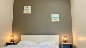 A bedroom with a bed, two bedside tables with lamps, and two wall-mounted paintings depicting beach scenes. - La Tranquille - House for 3 families (Saint-Denis-d'Oléron)