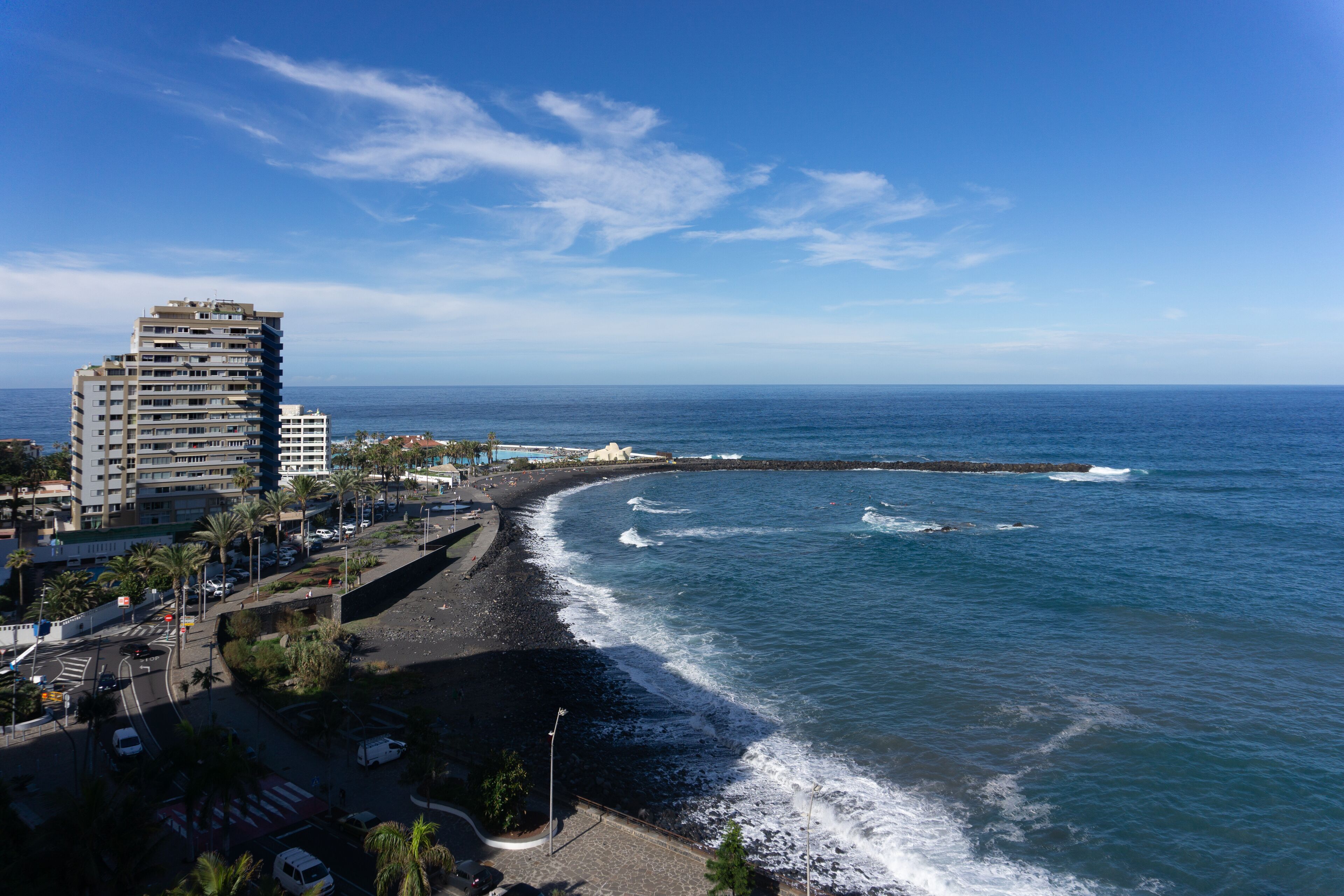 A coastal cityscape with a beachfront promenade, a tall hotel, and a rocky breakwater.