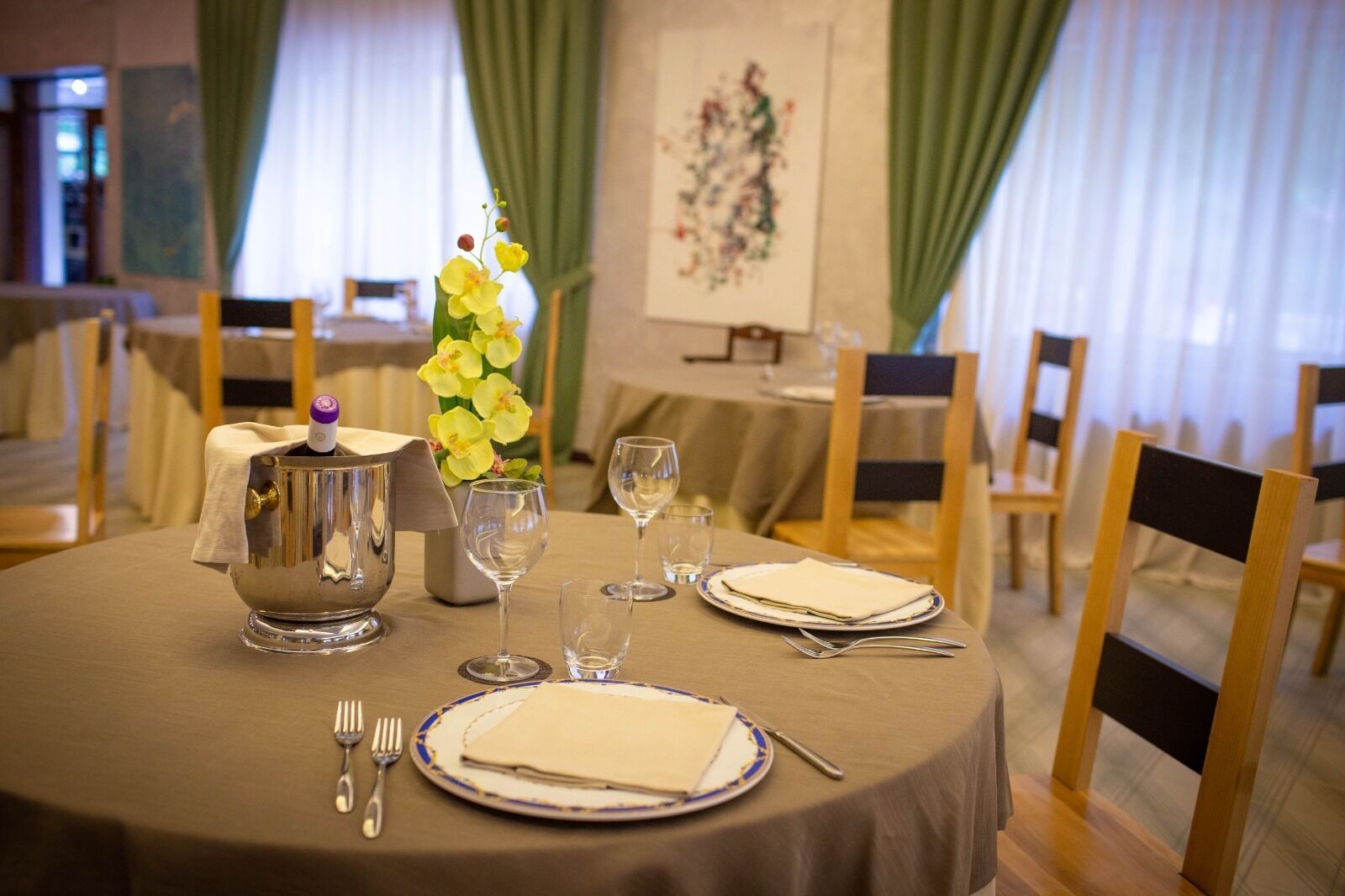 Mesa de comedor dispuesta con una jarra de plata, copas de vino y platos, rodeada de sillas en una habitación con cortinas y un cuadro en la pared.