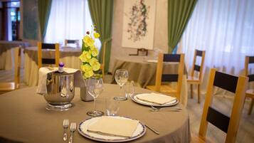 Mesa de comedor dispuesta con una jarra de plata, copas de vino y platos, rodeada de sillas en una habitación con cortinas y un cuadro en la pared.