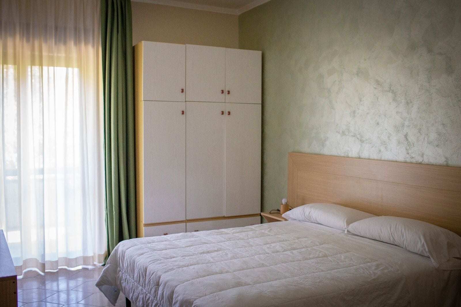 Ein Schlafzimmer mit Bett, Kleiderschrank, Nachttisch und einem Fenster mit Vorhängen.