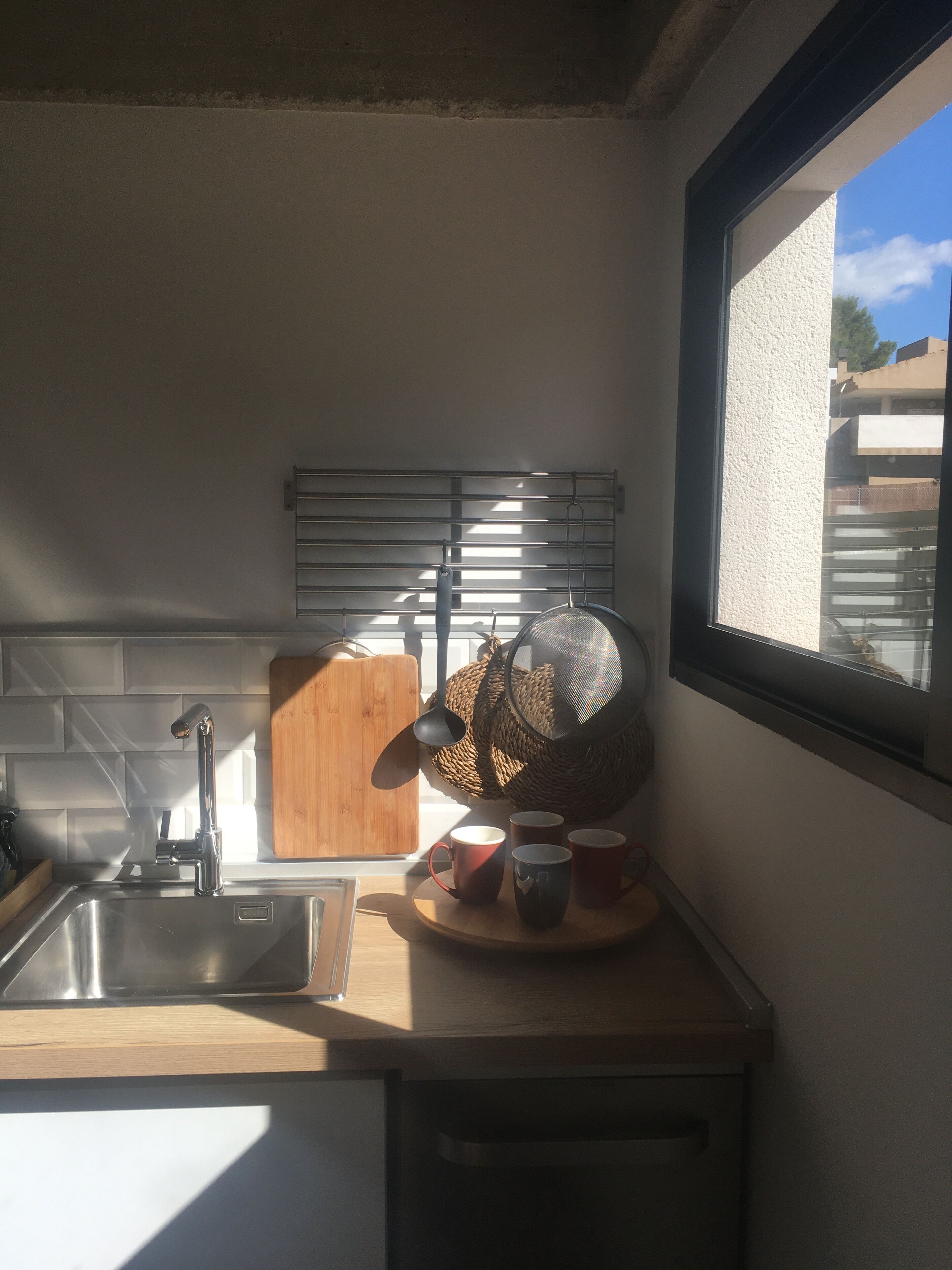 Een keuken met een raam, een spoelbak, een houten snijplank en een vergiet op het aanrecht.
