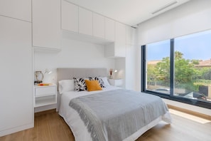 A modern bedroom with a large window, a bed with a grey blanket, white nightstands, and a white wardrobe.