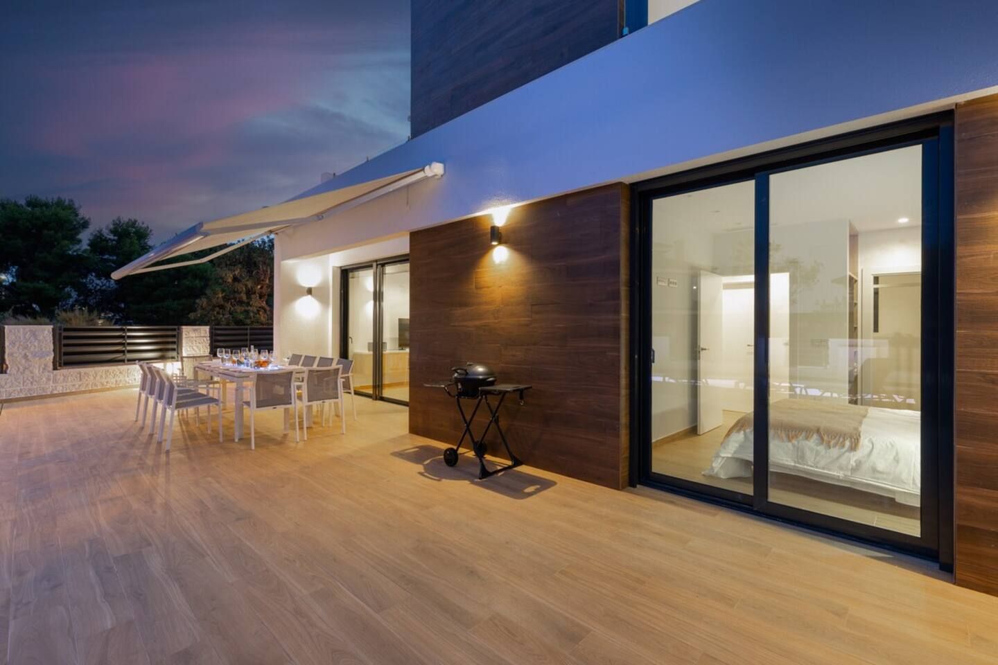 Een modern terras buiten met een eettafel en stoelen, een barbecue en een kamer met een bed, volledig omgeven door glas.