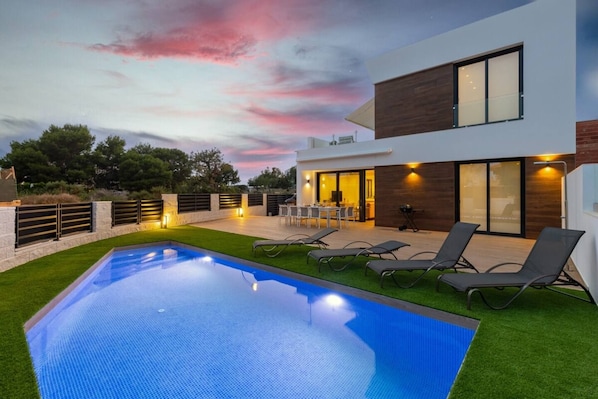 A modern house with a swimming pool, outdoor seating, and a sunset sky.