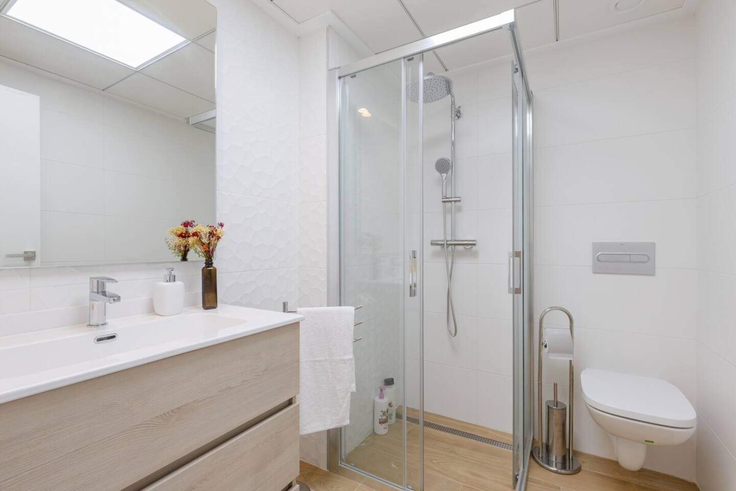 A modern bathroom with a glass shower enclosure, a white toilet, and a wooden vanity with a mirror and a vase of flowers.