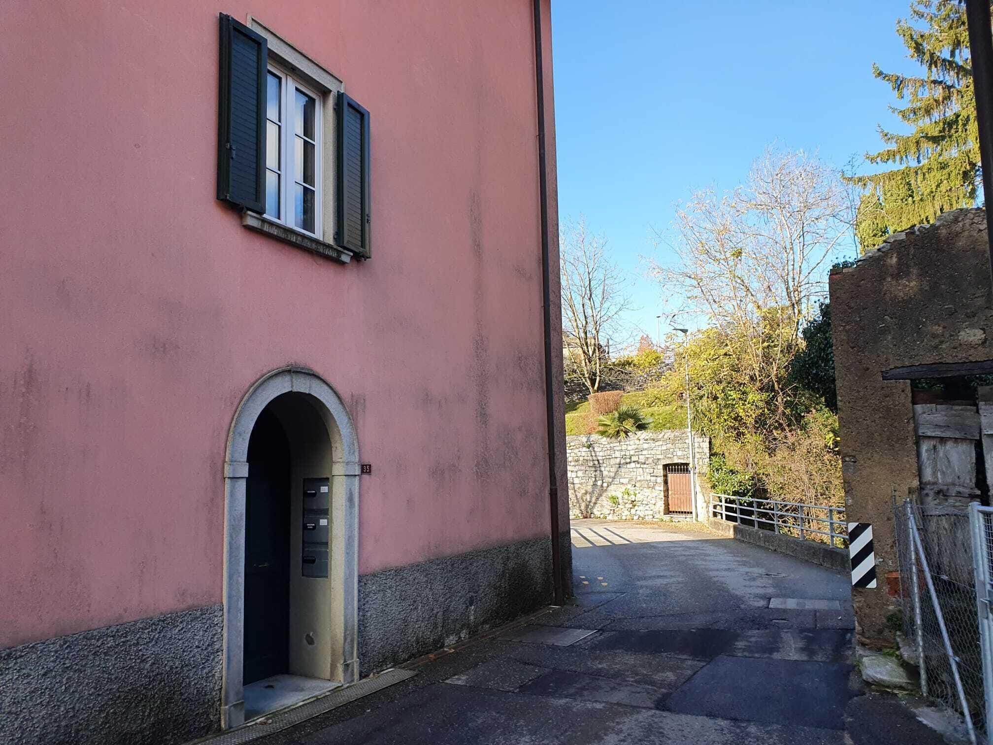 A narrow street with a pink building featuring a small arched entrance and a window with shutters.