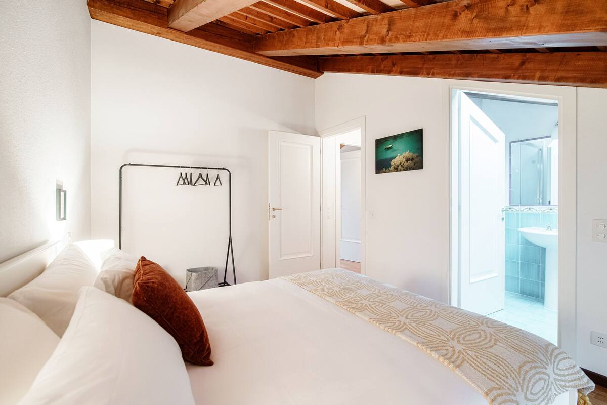 A bedroom with a large bed, a wooden ceiling, a white wall, a door, and a bathroom visible through an open door.