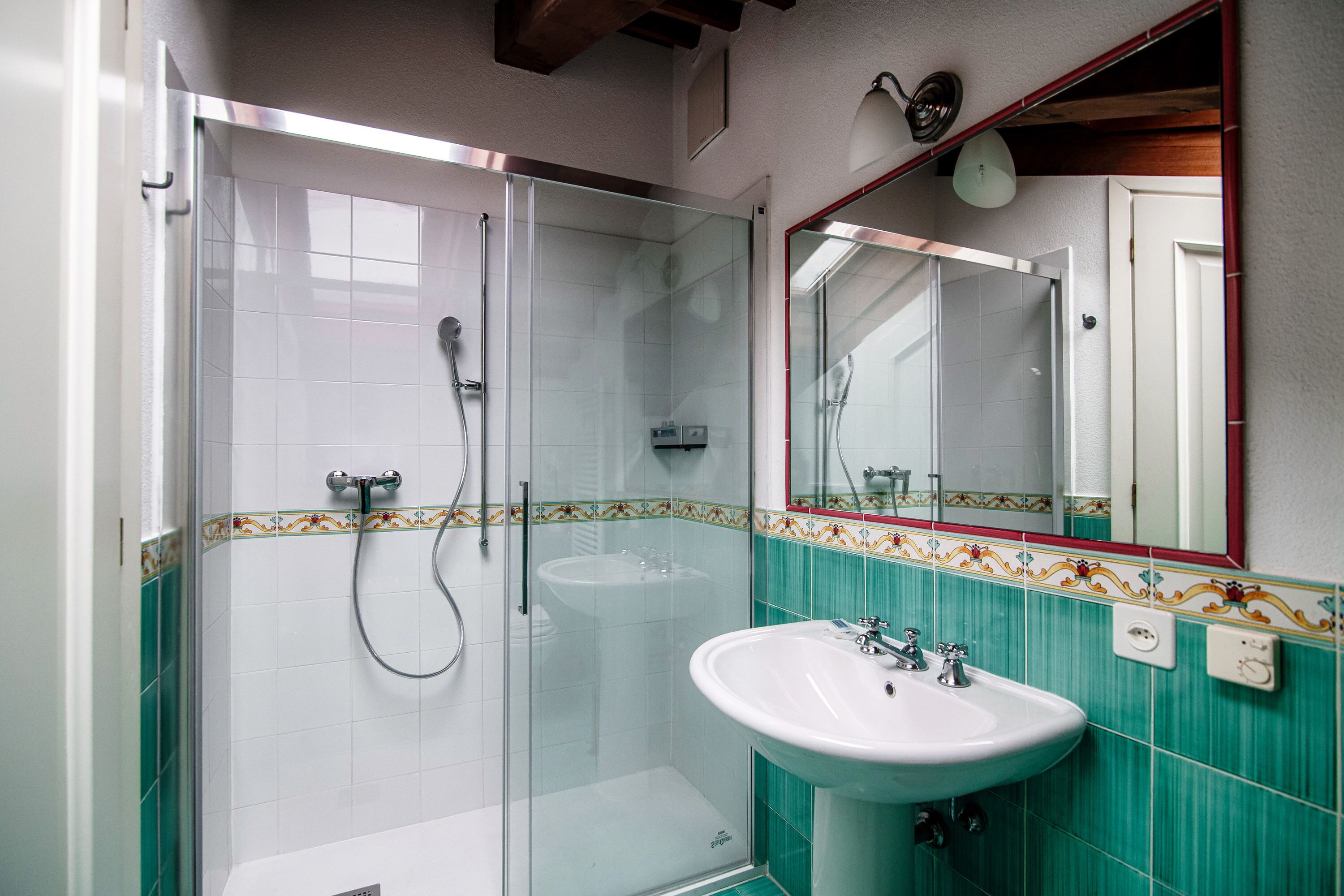 Ein Badezimmer mit einer gläsernen Duschkabine, einem weißen Waschbecken und einem Spiegel mit Zierrahmen.