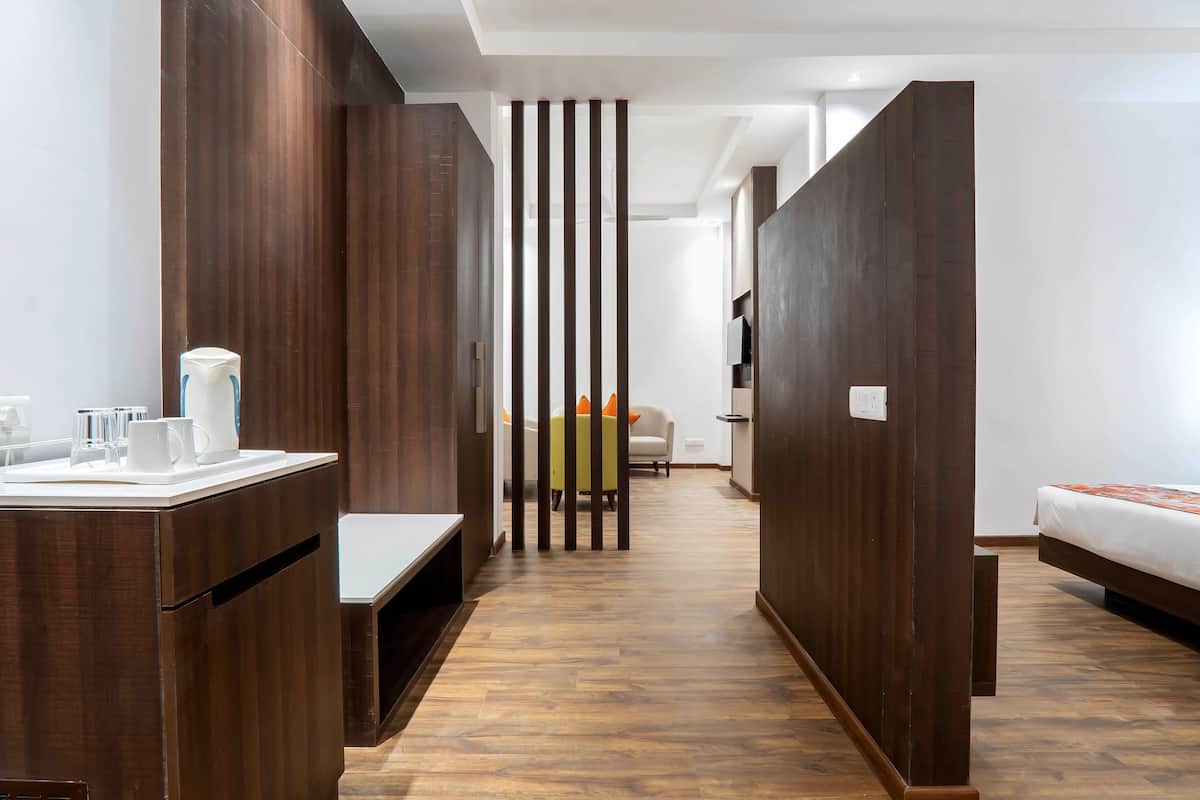 A modern hotel room with a wooden floor, a desk with a coffee maker, a bed, and a partition wall with vertical slats.