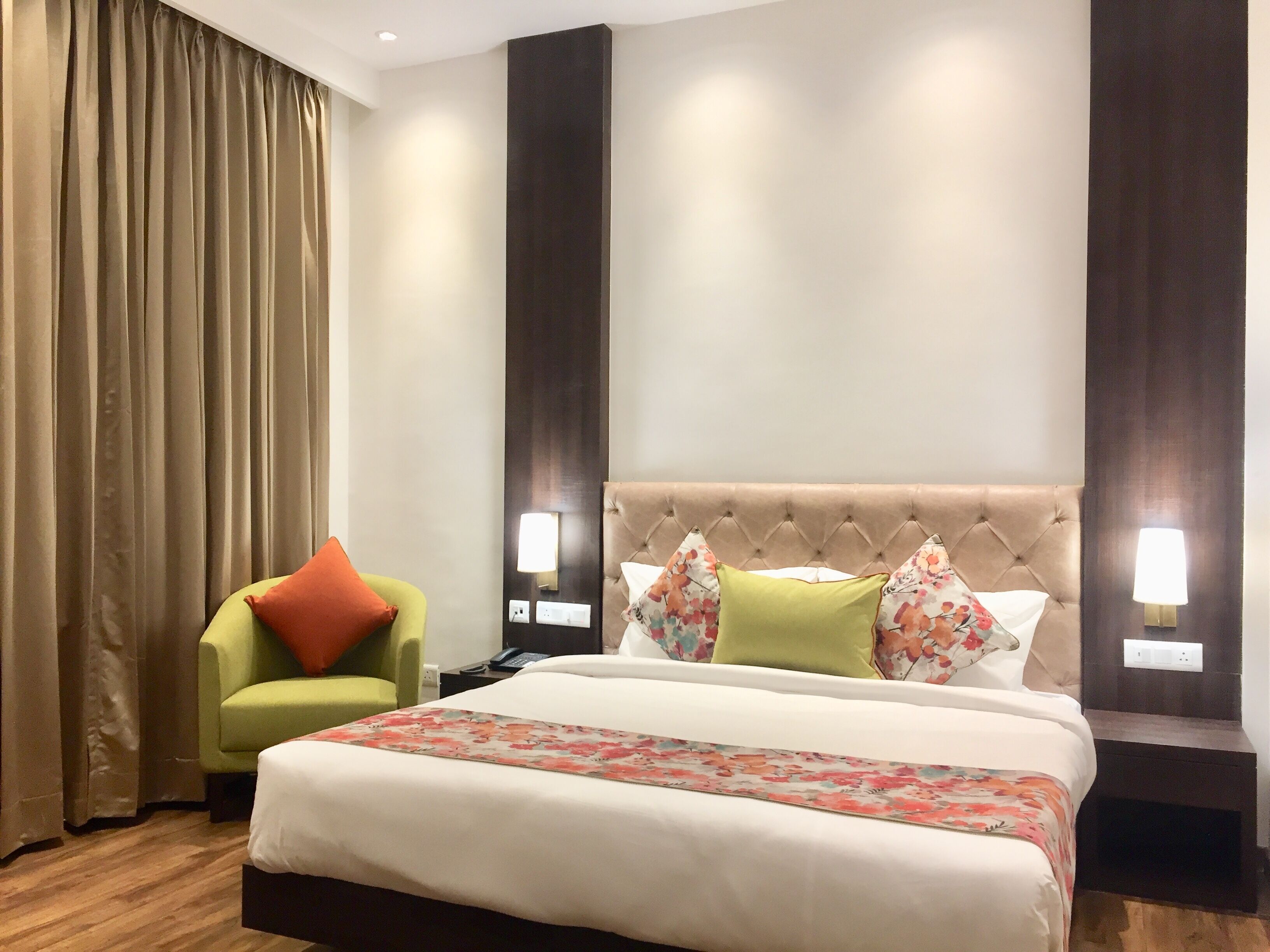 A hotel room with a bed, a green armchair, a large window with curtains, and wall-mounted lamps.