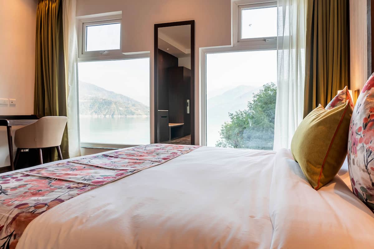 A hotel room with a bed, a desk, and a chair. There are two windows offering a view of the outdoors.