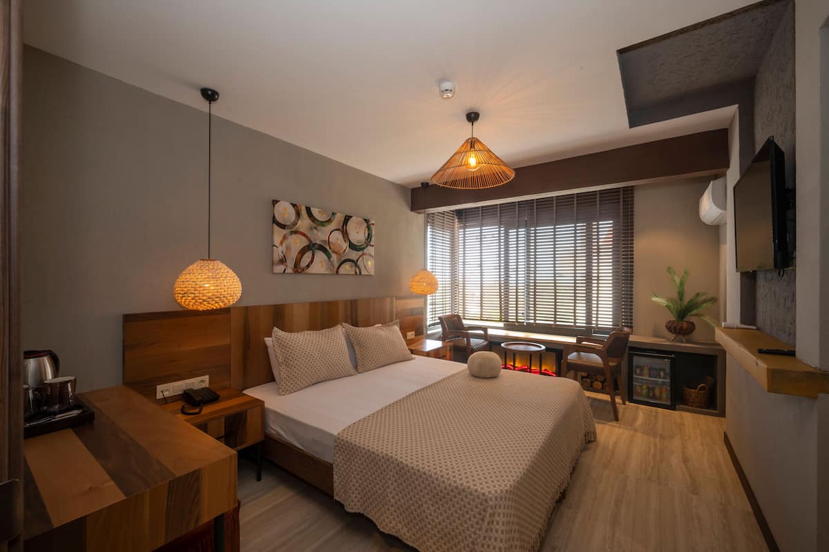 Une chambre d’hôtel moderne avec une tête de lit en bois, un grand lit recouvert d’une courtepointe à motifs, une table de chevet avec une lampe, un petit bureau avec une chaise, une fenêtre avec des stores et un tél éviseur fixé au mur.