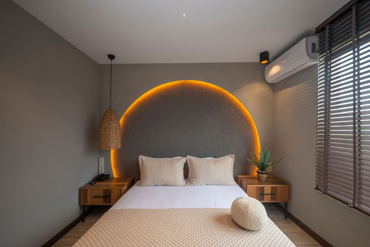 Une chambre avec un grand lit, un luminaire circulaire au-dessus du lit, une table de chevet avec une lampe et une fenêtre avec des stores.