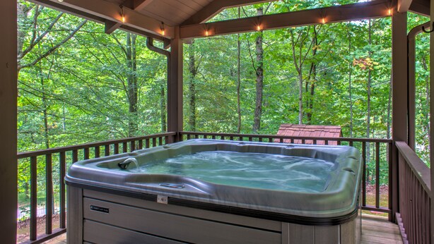 A hot tub in a covered outdoor area with a view of a forest. - Pisgah Pines | Cozy Log Cabin w/ Hot Tub & Fire Pit Near Asheville Airport (Arden)