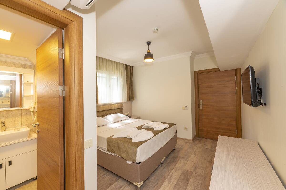 A bedroom with a bed, a wooden wardrobe, a television mounted on the wall, and a bathroom with a sink and mirror.