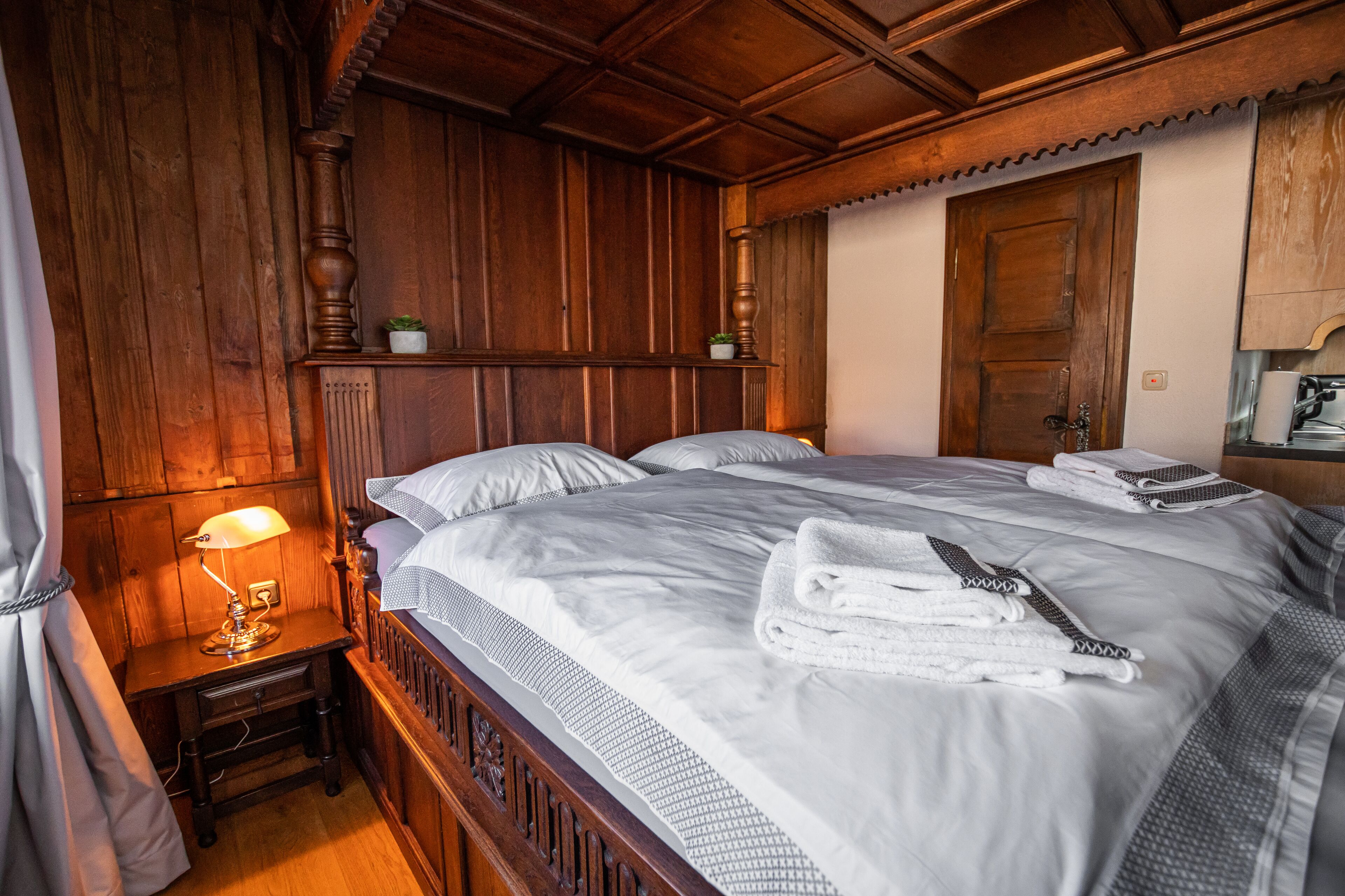 A wooden-paneled room with a large bed, a nightstand with a lamp, and a wooden door.