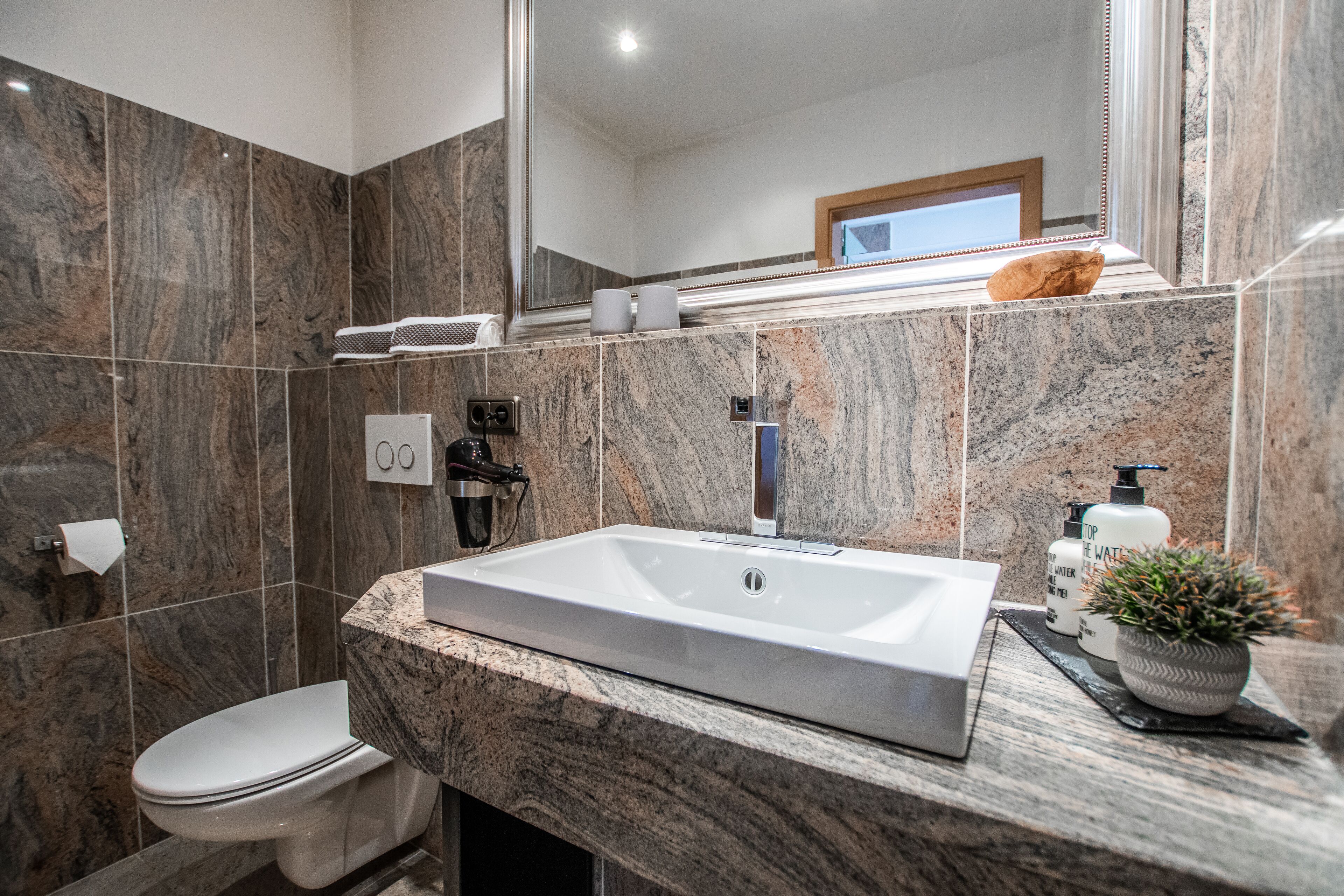 Uma casa de banho moderna com lavatório branco, bancada de mármore e um espelho grande.