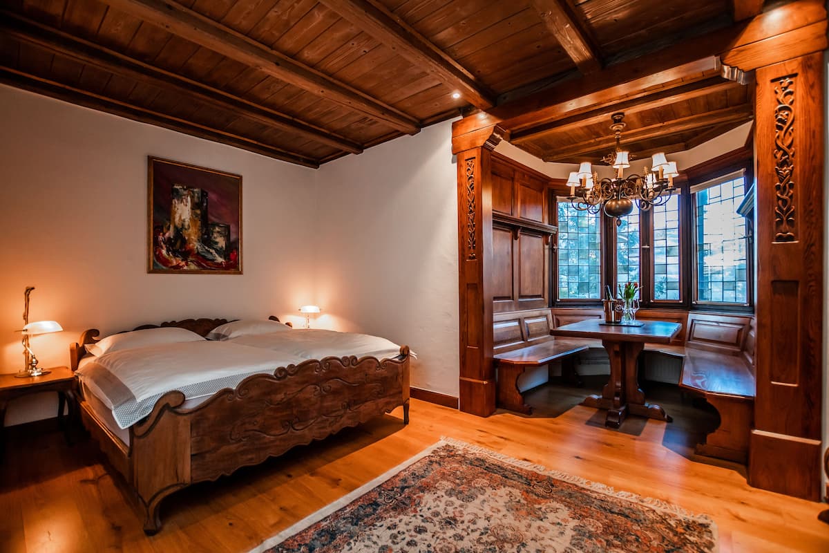 A bedroom with a wooden bed, a nightstand, a desk with a chair, and a window with a view.