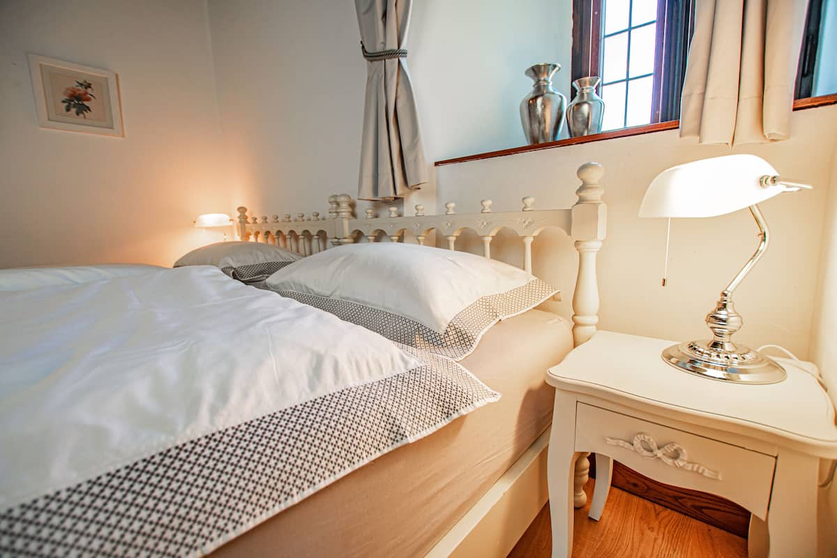 A neatly made bed with white linens and a patterned throw, a bedside table with a lamp, and a window with curtains.