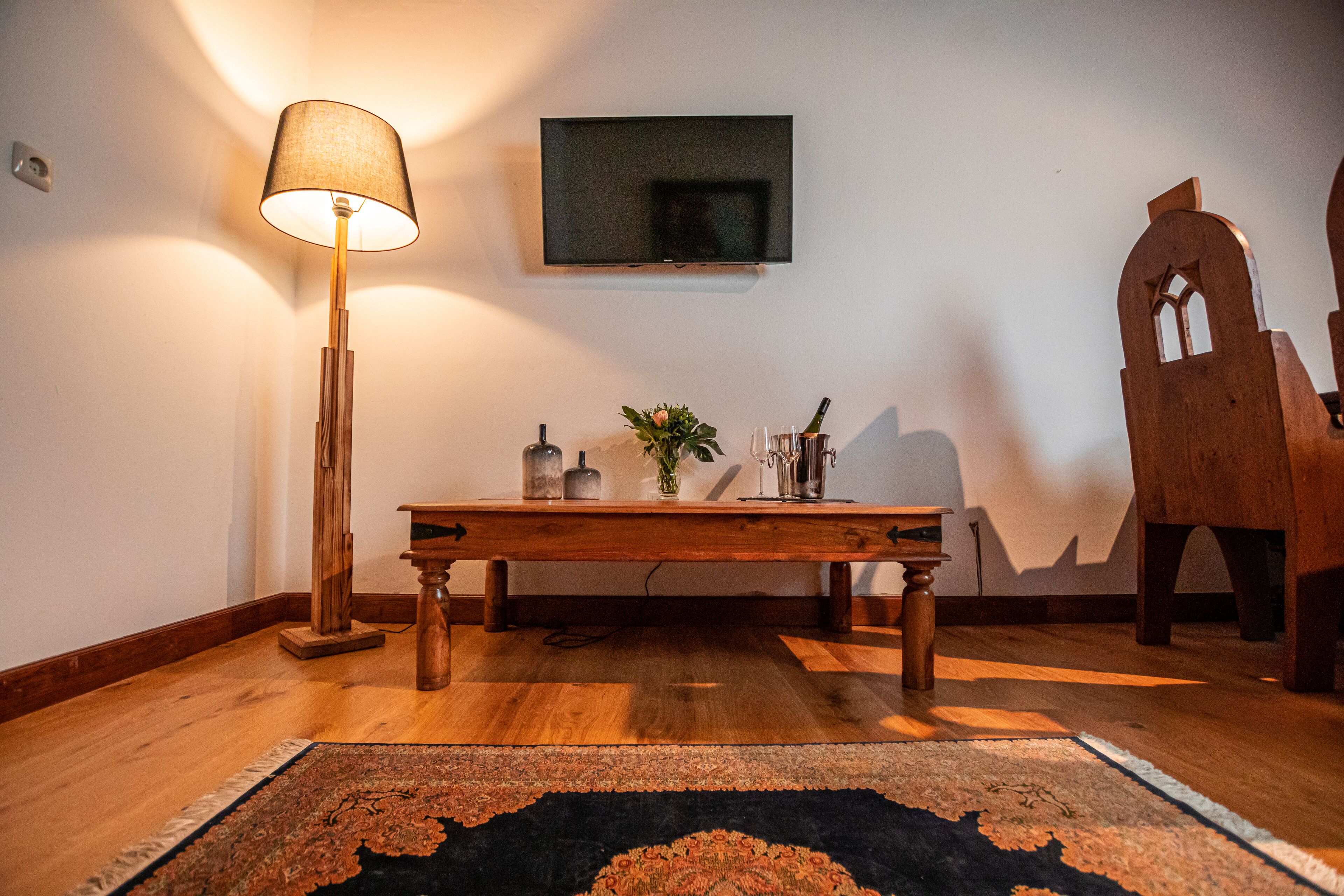 Una mesa de madera con una lámpara, un televisor de pantalla plana y una silla de madera decorativa en una habitación con piso de madera y un tapete estampado.
