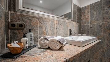 Un baño con encimera de mármol, un lavabo blanco y un espejo grande. En la encimera hay toallas, artículos de aseo y un cuenco de madera.