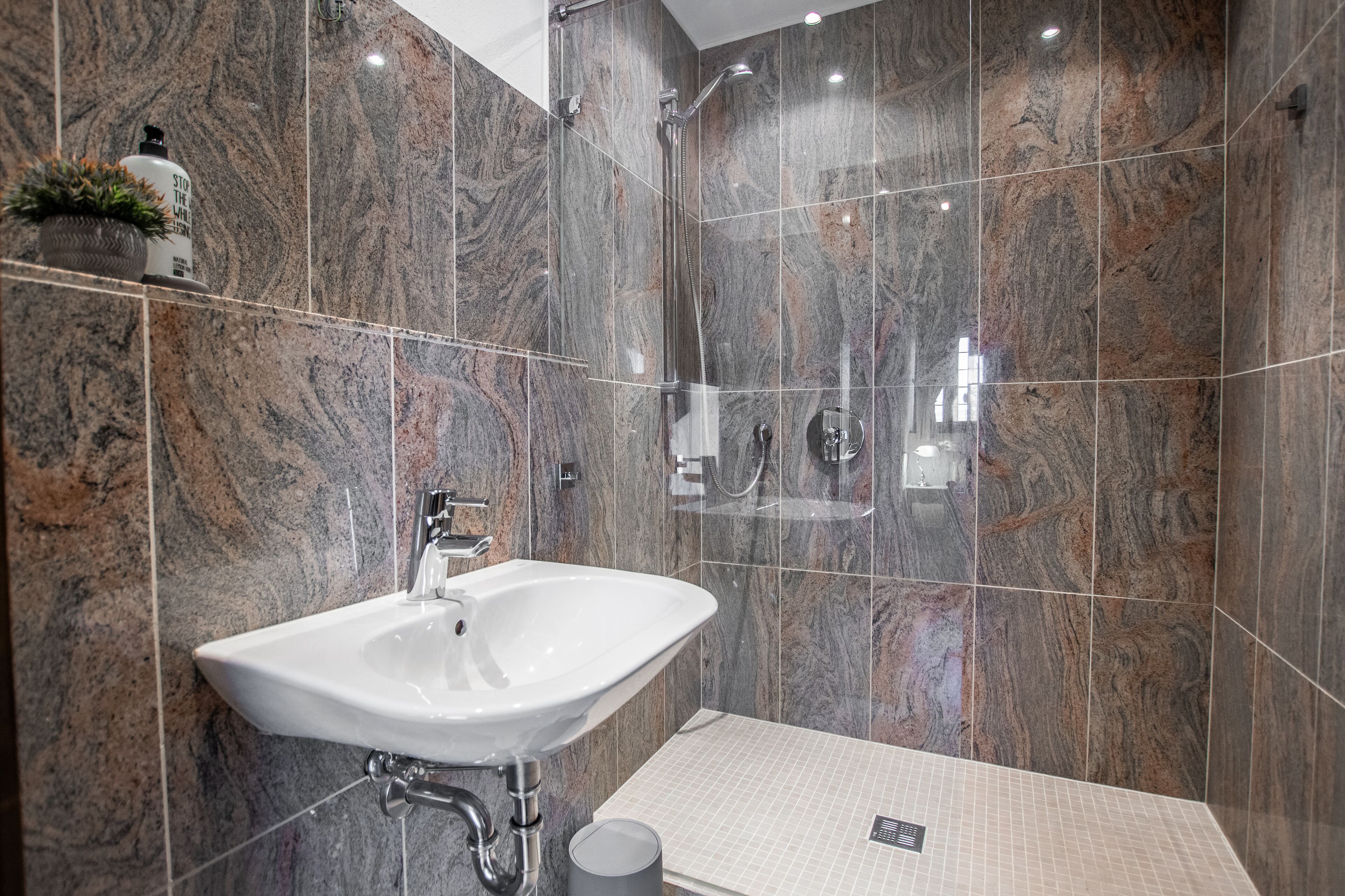 A modern bathroom with a marble wall, a white sink, a shower, and a small potted plant.
