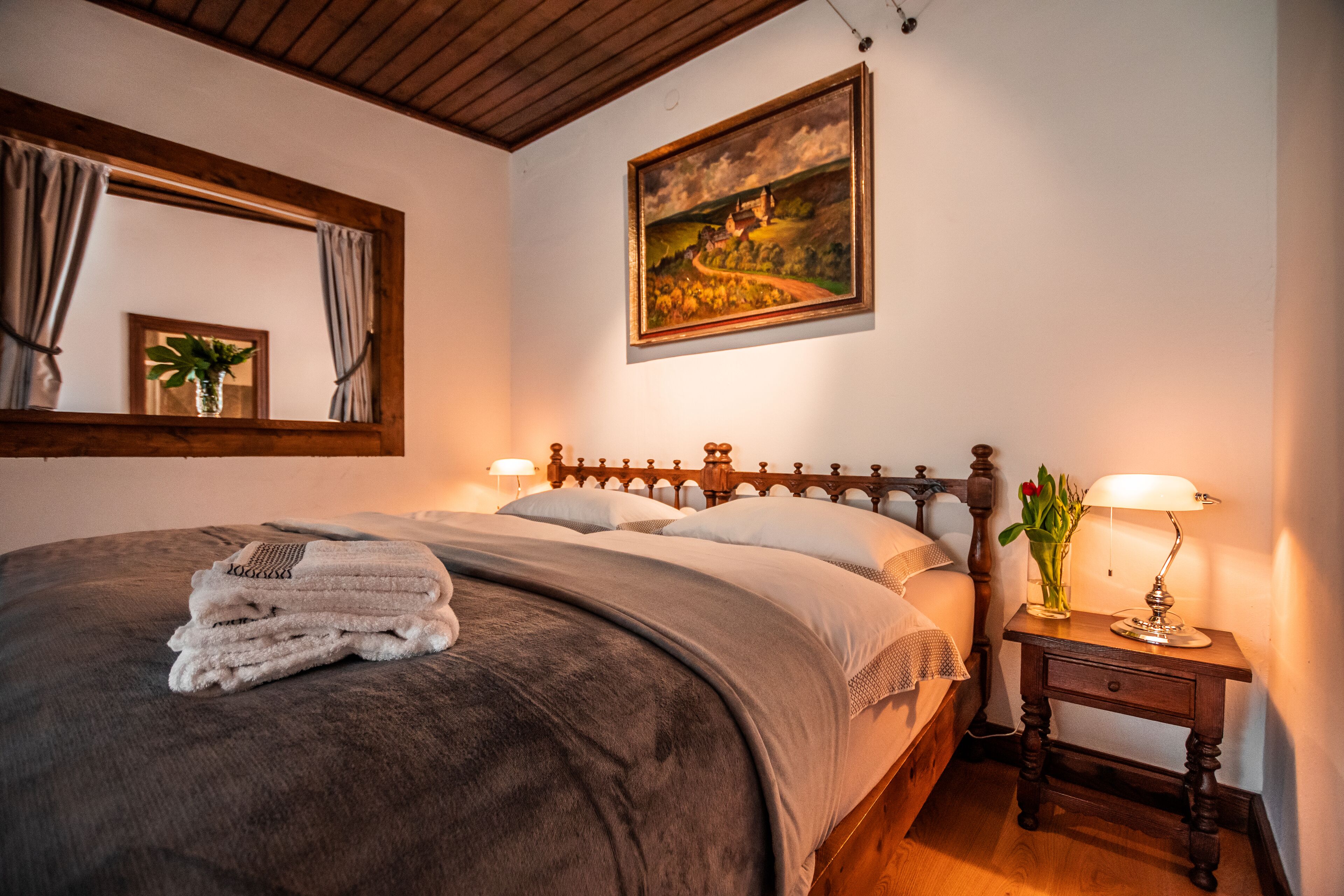 A neatly made bed with a gray blanket and a stack of white towels, a wooden nightstand with a lamp, a framed painting on the wall, and a mirror with curtains.