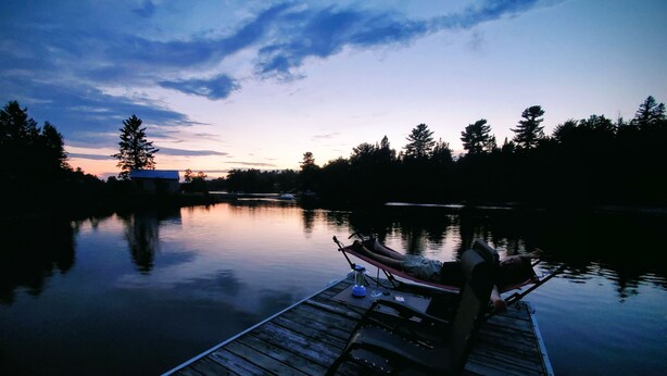 A wooden dock extending into calm water with a chair and a small table, surrounded by trees and a clear sky. - Getaway with Snowmobile, ATV, and boating access!! (Rockwood)