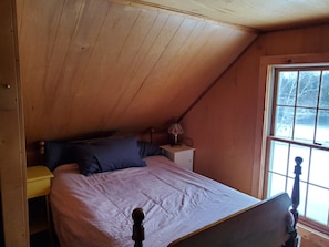 Un dormitorio con techo inclinado, cama con cabecera de madera, mesita de noche con lámpara y ventana con vistas a árboles.
